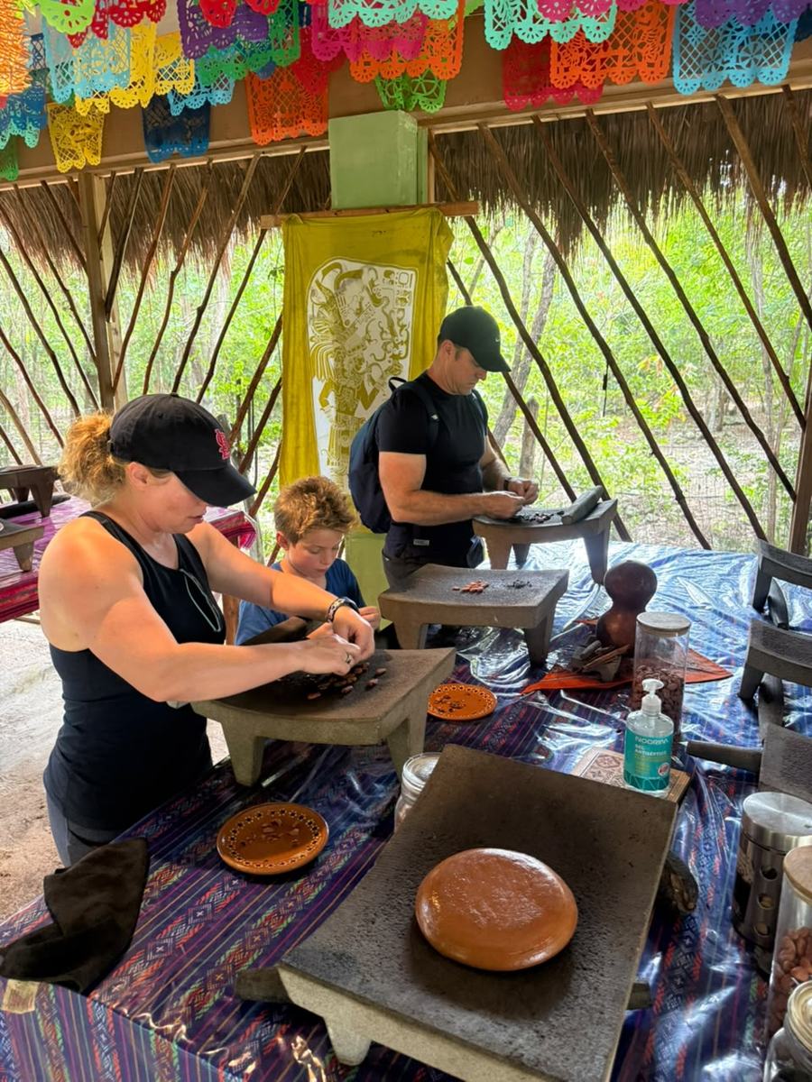 A family of three grinding cacao, a Maya banner behind them